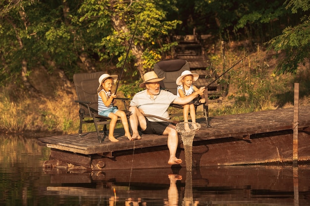 Симпатичные маленькие девочки и их дедушка ловят рыбу на озере или реке. Отдых на пирсе рядом с водой и лесом во время заката летнего дня. Понятие семьи, отдыха, детства, природы.