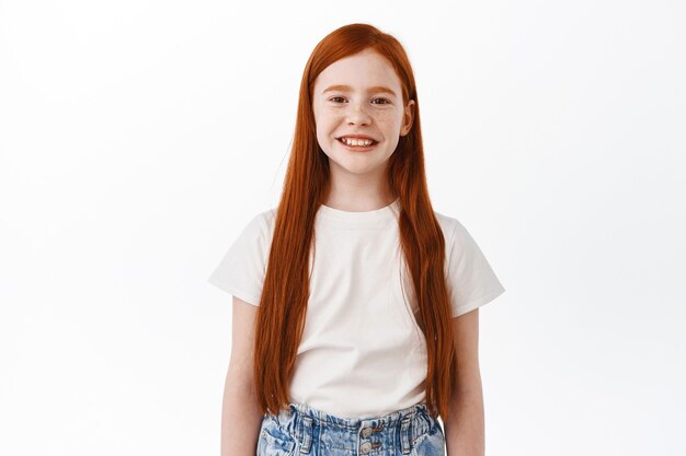 Милый маленький ребенок с длинными рыжими волосами улыбается и выглядит счастливым спереди, стоя над белой стеной
