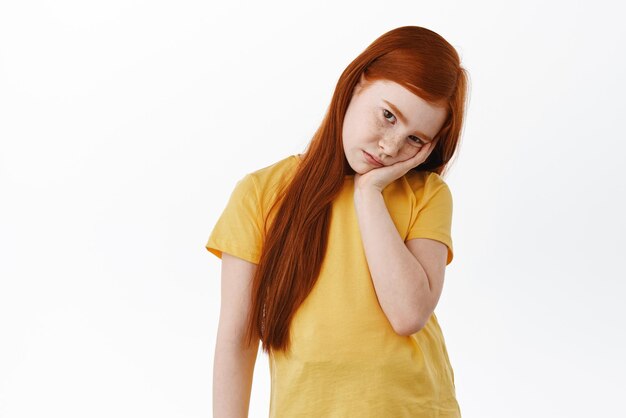 Cute little redhead girl feeling bored lean head on palm and stare indifferent and sad at camera Gloomy kid stare at something boring white background