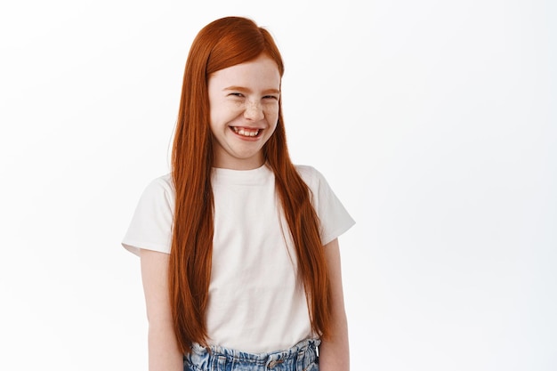 Милая маленькая рыжая девочка с длинными рыжими волосами и веснушками, смеясь и улыбаясь, хихикает над чем-то смешным, стоя на белом фоне