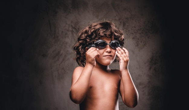 Cute little shirtless boy in swimming goggles posing in a studio. Isolated on a dark textured background.