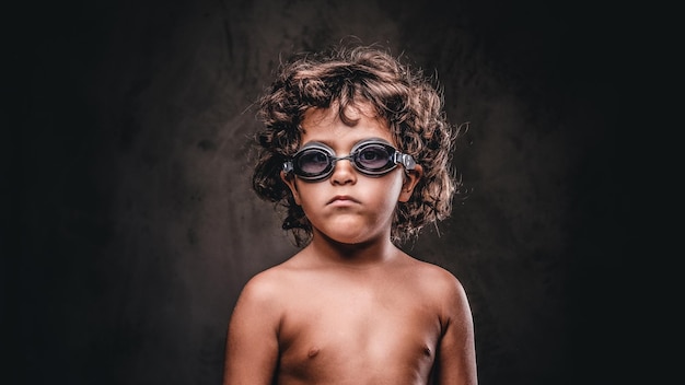 Cute little shirtless boy in swimming goggles posing in a studio. isolated on a dark textured background.