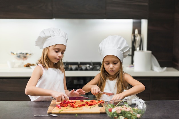 Cute little sisters cutting bell pepper on cutting board while preparing food