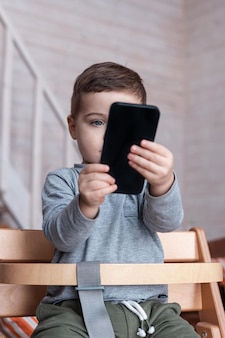 Симпатичный маленький мальчик, играющий со смартфоном, здоровый ребенок, касающийся мобильного телефона пальцами, выглядит ...