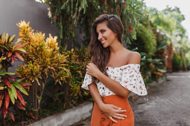 Cute long-haired woman in bright skirt and light blouse posing in tropical garden