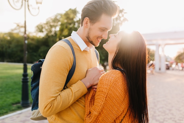 Cute long-haired woman in knitted orange sweater looking into boyfriend's eyes with love in sunny autumn day