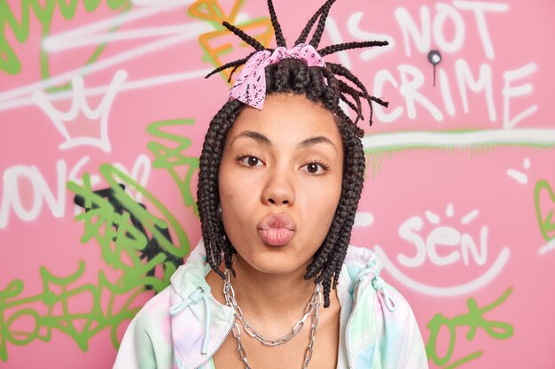 Cute lovely teenage girl keeps lips folded wants to kiss you has combed dreadlocks dressed in stylish clothes poses against colorful graffiti wall