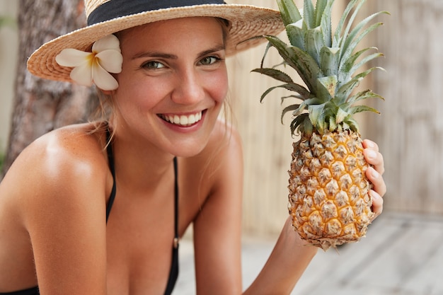 Cute lovely young woman wears bathing suit and straw hat, holds pineapple, happy to spend summer vacations in tropical country and eat exotic fruit. People, rest and healthy eating concept.