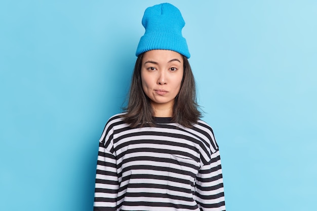 Cute millennial girl with dark hair has determined serious expression raises eyebrows makes assumptions looks directly, wears hat casual striped jumper 