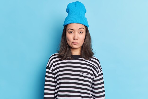 Cute millennial girl with dark hair has determined serious expression raises eyebrows makes assumptions looks directly, wears hat casual striped jumper 