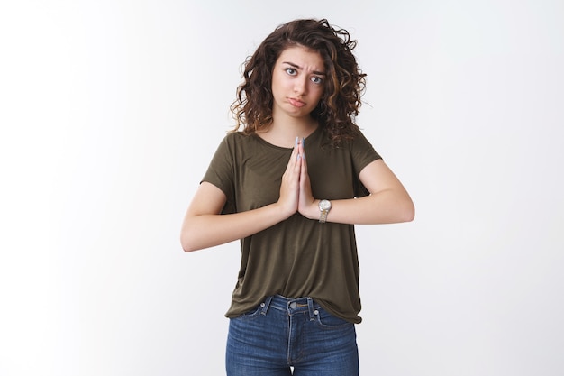 Cute miserable-looking young caucasian curly-haired woman sulking making pitty sad grimace press palms together pray supplicating begging help hopefully looking whining wanna favor say please
