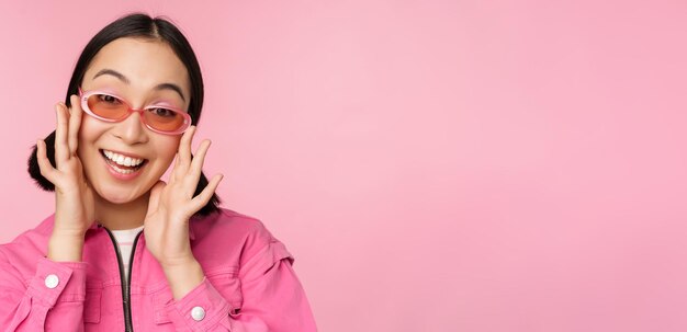 Cute modern japanese girl in sunglasses smiling and looking happy posing against pink background in stylish clothing