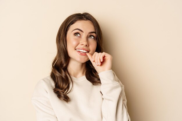 Cute modern woman thinking daydreaming and smiling looking up thoughtful standing over beige background