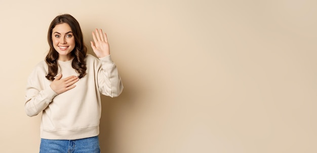 Cute modest girl smiling raising finger introduce herself saying hello standing over beige background