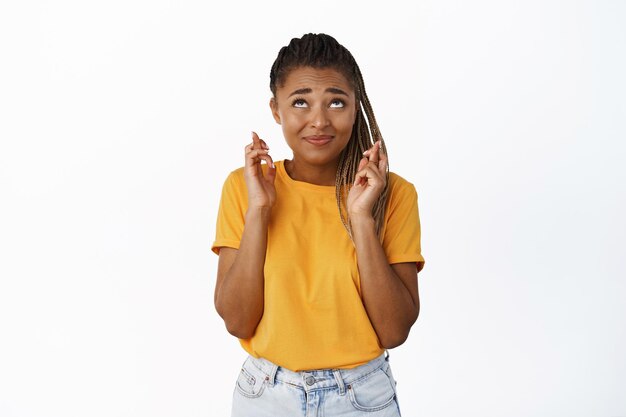Cute nervous Black girl student wishing looking up with fingers crossed praying standing over white background