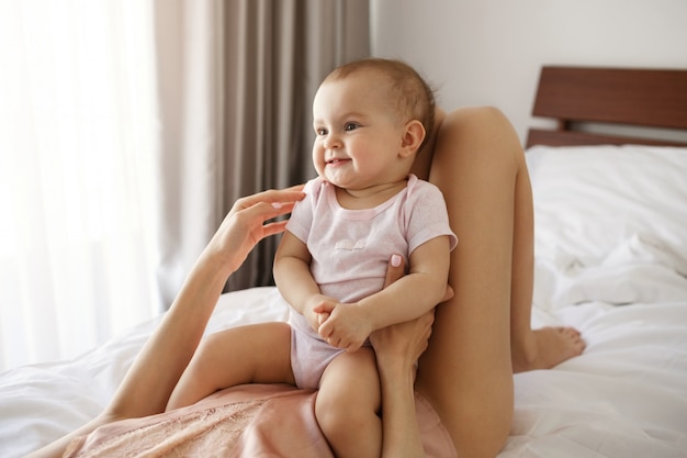 Cute nice baby daughter sitting on her young mom lying on bed smiling at home.