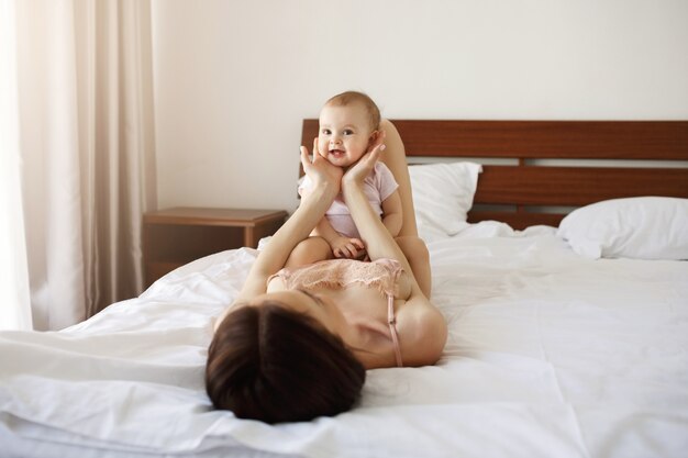 自宅で笑顔のベッドに横になっている若いお母さんの上に座ってかわいい素敵な赤ちゃん娘。