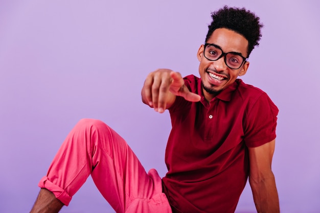 Cute optimistic guy pointing finger and laughing. Indoor shot of stylish african young man sitting.