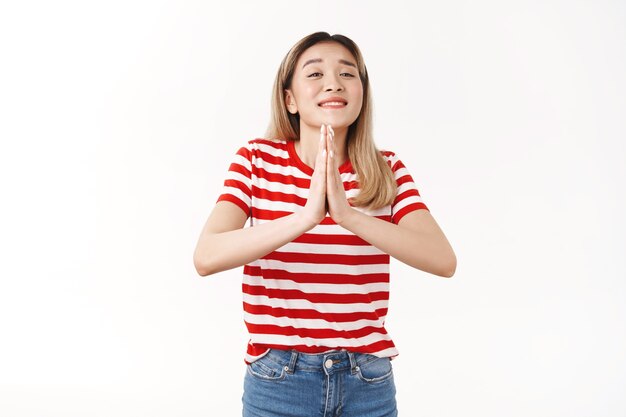 Cute optimistic silly blond asian girl begging friend lend hand do favour press palms together pray gesture close eyes smiling broadly pleading, making wish supplicating positive mood