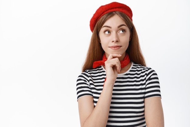 Foto gratuita carina ragazza pensierosa sorride e guarda da parte con interesse, pensando a qualcosa, in piedi contro il muro bianco