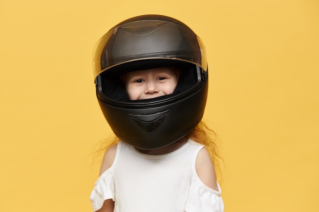 彼女の父から取られた黒いオートバイのヘルメットをかぶっているかわいい遊び心のある少女。笑顔で、保護モーター機器で隔離ポーズをとって面白い女児