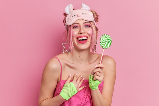 Cute positive Caucasian woman feels grateful for compliment holds candy smiles broadly wears soft sleepmask and dress 