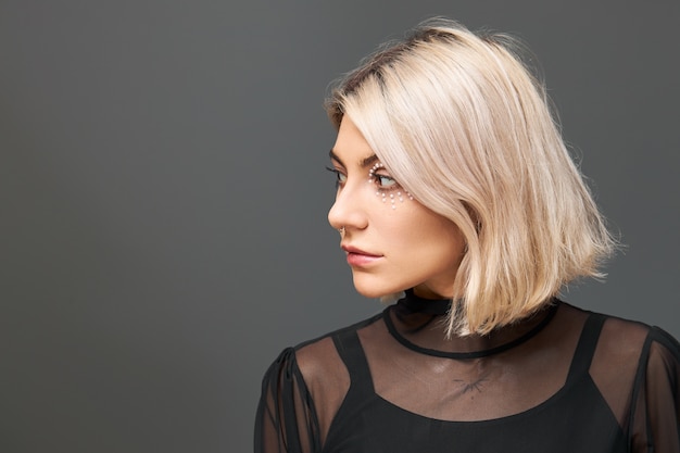 Cute positive young Caucasian female wearing trendy black top and transparent dress having dreamy pensive facial expression, making plans for Christmas holidays, looking away and smiling joyfully