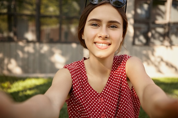 Cute pretty girl with broad charming smile taking selfie using electronic gadget.