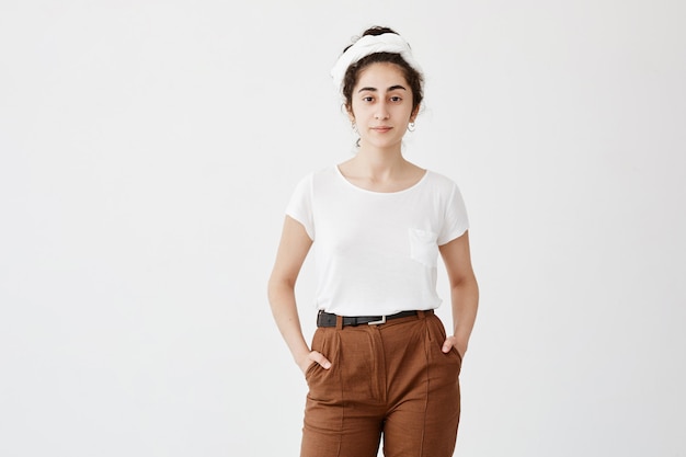 Cute pretty teenage girl wears loose white t-shirt and brown trousers, holds her hands in pocket, feels enjoyment. Pleasant-looking girl posing against white wall