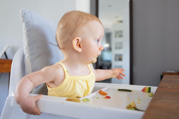쟁반에 지저분한 음식과 함께 유아용 의자에 앉아 멀리보고 얼굴에 녹색 퓌레 얼룩이있는 귀여운 빨간 머리 아기 소녀. 측면보기. 수유 과정 또는 육아 개념