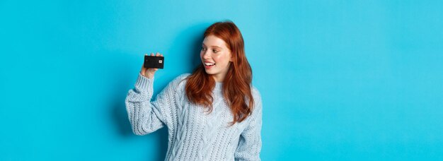 Carina ragazza rossa in maglione che mostra la carta di credito sorridendo alla telecamera in piedi su sfondo blu