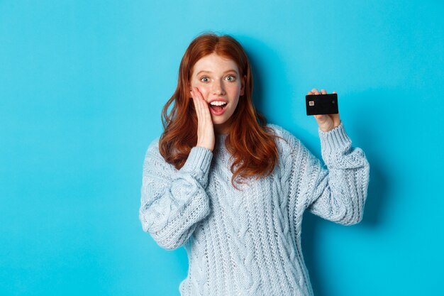 신용 카드를 보여주는 스웨터에 귀여운 빨간 머리 소녀, 카메라에 미소, 파란색 배경 위에 서