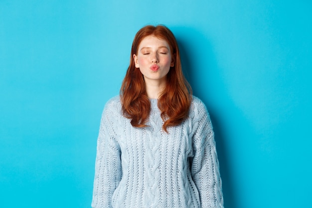 青い背景のセーターに立って、キス、パッカーの唇と目を閉じるのを待っているかわいい赤毛の十代の少女。