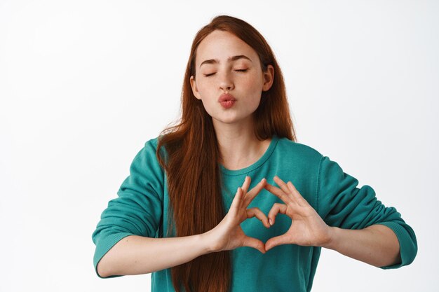 Cute redhead woman pucker lips, kissing and showing heart I love you sign, like you, feeling tender and romantic, standing in green blouse against white background