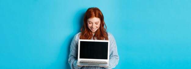 Free photo cute redhead woman in sweater showing and looking at laptop screen with pleased smile demonstrating