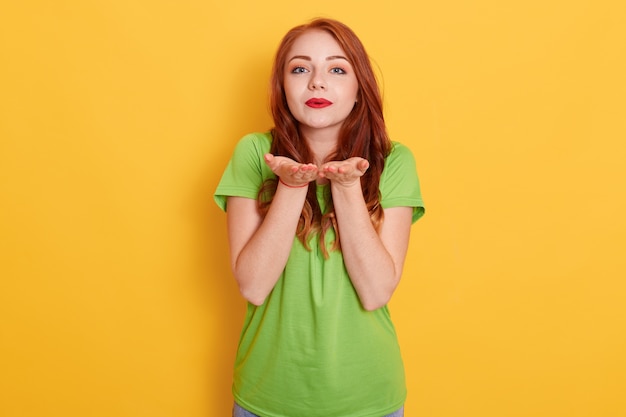 Cute romantic red haired woman standing and sending amorous air kiss to camera, demonstrating affection, wearing green t shirt