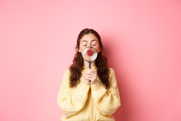 Cute and romantic young woman magnify her lips with magnifying glass, making kissing face, standing with closed eyes against pink background