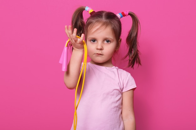 Бесплатное фото Милая, грустная девушка держит в руке скакалку. маленький ребенок хочет играть с кем-то. очаровательны малыш с пони хвостами и разноцветными резинки для волос, носит майку на розе.
