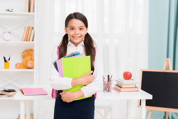 Cute schoolgirl in uniform holding notepads in classroom