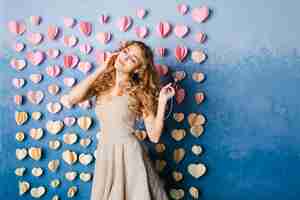 Free photo cute sexy slim girl with blond curly hair standing in a studio with blue background and listenign to music on earphones. she smiles and looks sexy.