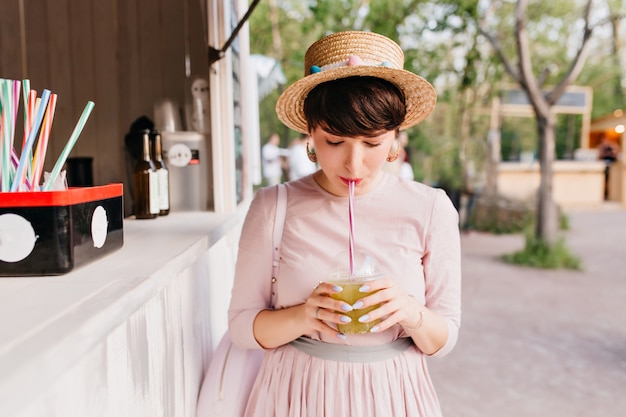 Cute short-haired young lady with elegant purple manicure drinking green cocktail standing near the snack-bar