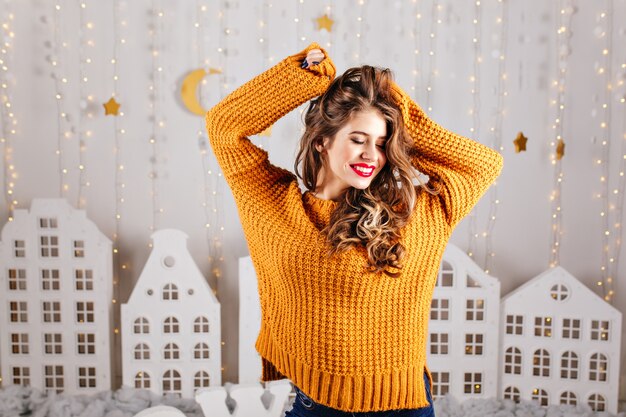 Cute, shy girl modestly smiles and poses with raised arms in cozy interior decorated for New Year