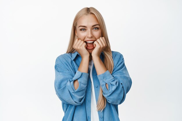 Cute and silly blond girl squeeze her cheeks and smiling looking amused at camera checking out something cool standing over white background