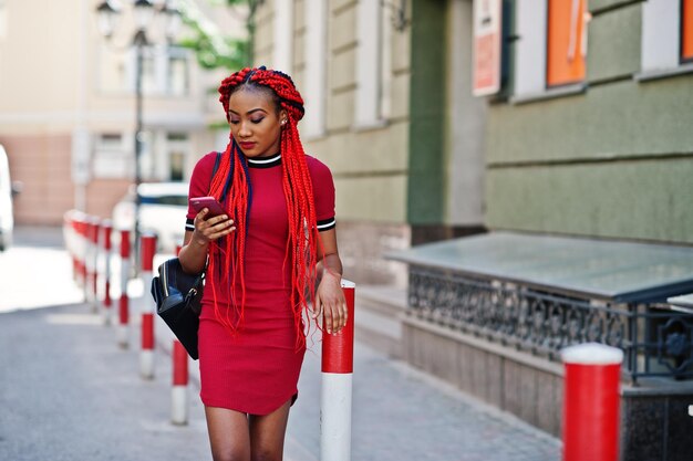 드레드락과 배낭을 메고 빨간 드레스를 입은 귀엽고 날씬한 아프리카계 미국인 소녀가 야외에서 포즈를 취하고 거리의 세련된 블랙 모델에서 휴대전화를 보고 있습니다