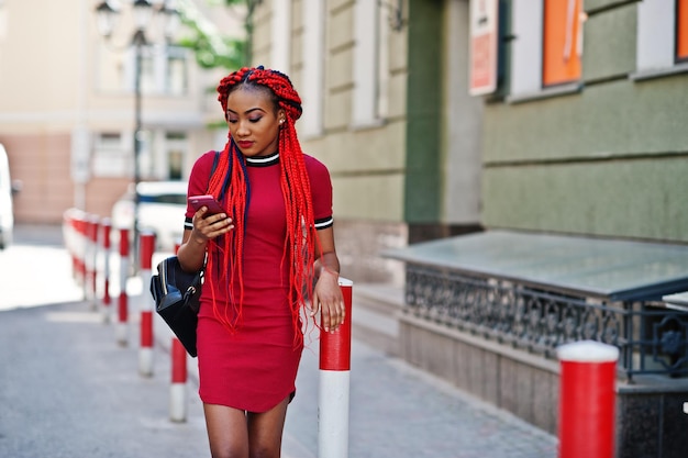 Симпатичная и стройная африканская американка в красном платье с дредами и рюкзаком позирует на улице и смотрит на мобильный телефон на улице Стильная черная модель
