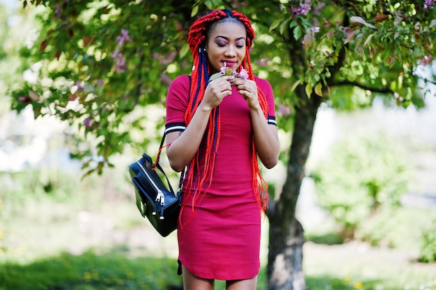 春の公園で屋外でポーズをとったドレッドヘアと赤いドレスのキュートでスリムなアフリカ系アメリカ人の女の子スタイリッシュな黒のモデル