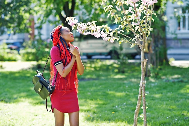 봄 공원에서 야외 포즈를 취한 드레드락이 있는 빨간 드레스를 입은 귀엽고 날씬한 아프리카계 미국인 소녀 세련된 블랙 모델