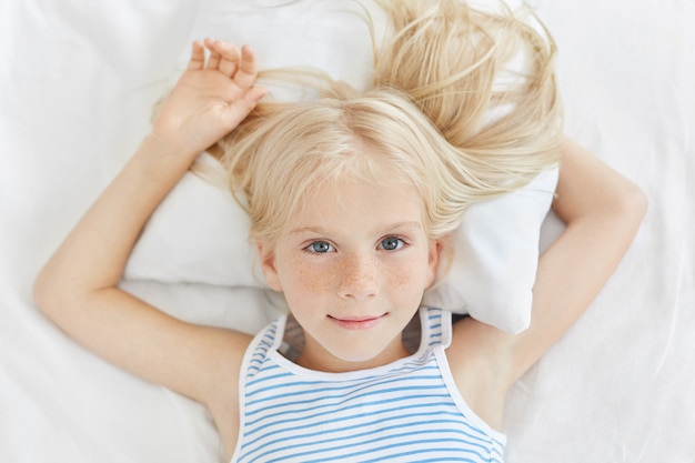 Cute small girl resting on white bed, looking with blue eyes. Dreamy blue eyed blonde girl in comfortable bedroom