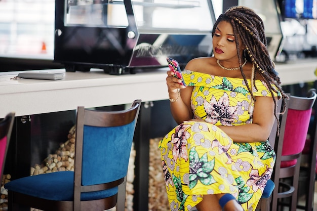 Бесплатное фото Милая африканская американка маленького роста с дредами в цветном желтом платье сидит в кафе в торговом центре и смотрит на мобильный телефон