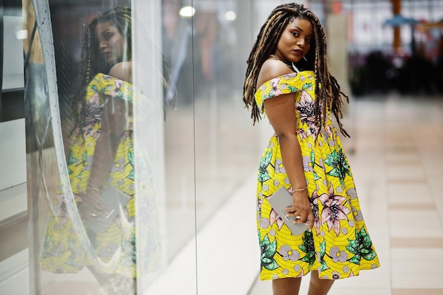 Симпатичная африканская американка маленького роста с дредами в цветном желтом платье, стоящая у витрины магазина в торговом центре